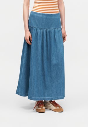 Žena v modré džínové maxi sukni, kterou doplňuje béžová a hnědá obuv a oranžovo-bílé pruhované tílko bez rukávů.