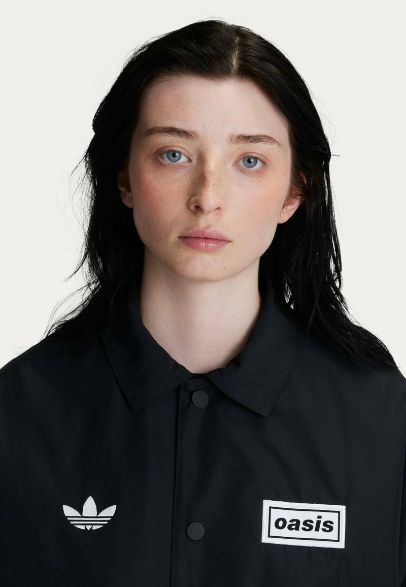 Black windbreaker with a collared neckline, featuring white "oasis" emblem and Adidas logo on the left chest. Smooth texture, button closure.