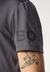 Nahaufnahme eines Arms mit einem Tattoo eines Krokodils, der ein graues Hemd mit aufgestickten schwarzen Buchstaben "BOR" trägt.