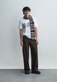 Άσπρο βαμβακερό t-shirt με γραφικό ζιγκ-ζαγκ σε μπλε και γκρι, καφέ παντελόνι με φαρδιά πόδια και φουλάρι με σχέδιο σε καφέ, κρεμ και σκούρο μπλε.