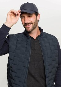 Mornarsko prešito brezrokavnik z visokim ovratnikom, v kombinaciji s temnim polo majico. Model nosi mornarsko kapo in jo prilagaja z eno roko, medtem ko se smeška.