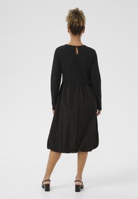 Crna haljina dugih rukava sa prorezom na leđima. Napravljena od teksturiranog materijala, s pojasom na naboranje i lepršavom suknjom. Čizme su vidljive.