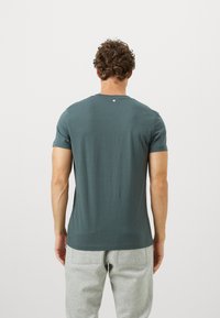 Άνδρας με καστανά σγουρά μαλλιά, που φοράει ένα σκούρο πράσινο κοντομάνικο μπλουζάκι και ανοιχτό γκρι παντελόνι, σ�τέκεται με την πλάτη προς την κάμερα.