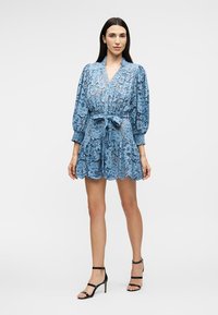 Woman wearing a blue floral lace dress with puffed sleeves and a tied waist, paired with black high-heeled sandals, standing against a white background.