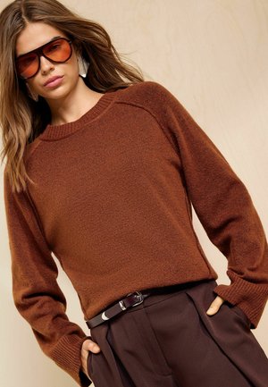 Frau trägt braunen Strickpullover, dunkelbraune Hose mit Gürtel, große getönte Sonnenbrille und auffällige Ohrringe, die Hände in den Taschen.