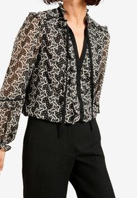 Blusa in bianco e nero con un motivo floreale, tessuto trasparente, colletto arricciato e chiusura con bottoni. Maniche lunghe con polsini elastici.