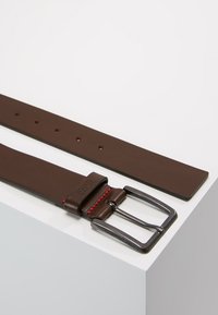 Bruine leren riem met een zilverkleurige metalen gesp, voorzien van een rode accent en meerdere gaatjes voor aanpassing. Gladde textuur en rechthoekige vorm.
