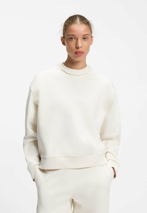 Jeune femme à la peau claire et aux cheveux tirés en arrière, portant un sweat-shirt blanc uni à manches longues et un pantalon assorti, les mains dans les poches, fond neutre.