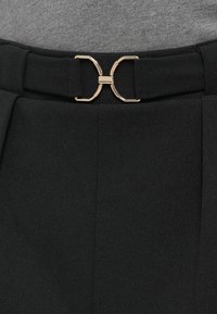 Μαύρη υφασμάτινη ζώνη με αγκράφα σε χρυσαφί απόχρωση και μοναδικό γεωμετρικό σχέδιο. Η υφή είναι λεία με διακριτικό μοτίβο.