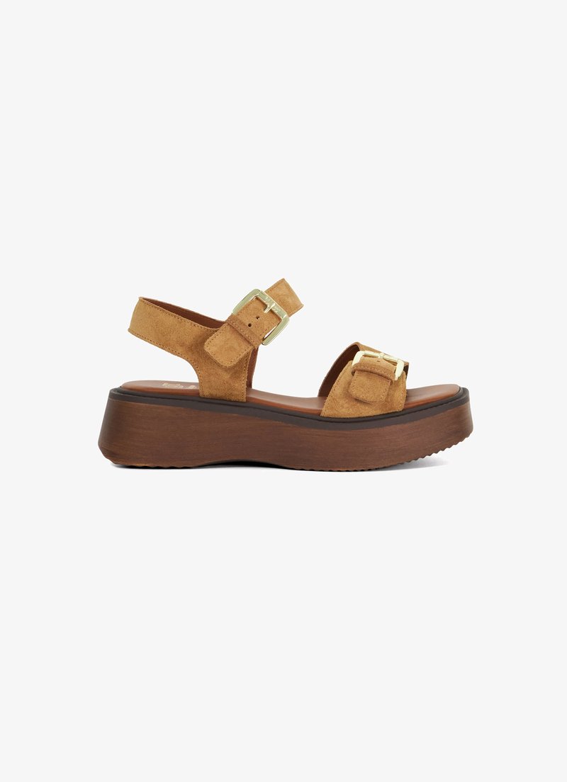 Tan suede platform sandals with adjustable ankle strap, buckle accents, and wooden textured sole. Flat heel with a slight curve.