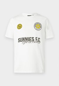 Bílé bavlněné tričko s krátkými rukávy. Obsahuje dva kulaté nášivky, jeden žlutý a jeden černý, a černý text „SUNNIES FC“ a „Lyle & Scott“.