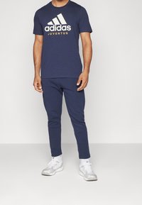 Mężczyzna ubrany w granatową koszulkę Adidas Juventus, dopasowane spodnie dresowe, białe sneakersy Adidas oraz białe skarpetki, stojący na jednolitym tle.