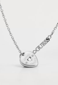 Silver-tone necklace with a heart-shaped pendant featuring "GUESS AMORE" engraving, centered rhinestone, and an attached logo accent.