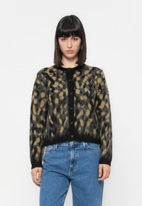 Mulher com cabelos lisos e escuros, vestindo um cardigan de estampa de oncinha abotoado e jeans azuis, em frente a um fundo branco simples.