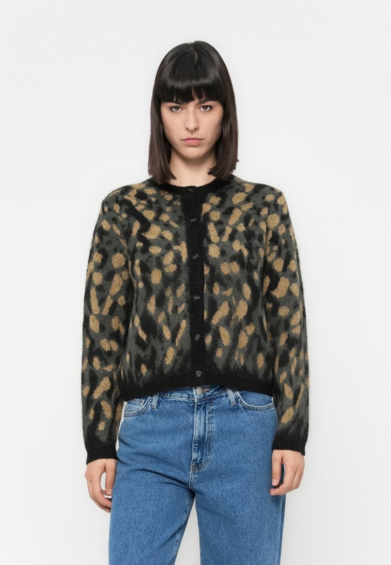 Mulher com cabelos lisos e escuros, vestindo um cardigan de estampa de oncinha abotoado e jeans azuis, em frente a um fundo branco simples.