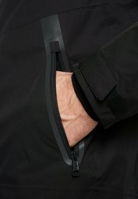 Giubbotto impermeabile nero con tasca con zip caratterizzata da una zip grigia a contrasto. Include dettagli in velcro sui polsini e una texture liscia.