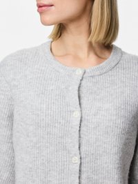 Lysegrå knappet cardigan laget av mykt, teksturert stoff med ribbestrikket mønster og rund hals. Har hvite knapper langs fronten.