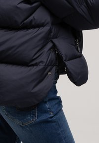 Granatowa kurtka puchowa z pikowanymi sekcjami, widocznym zamkiem błyskawicznym, zatrzaskami i elastycznymi mankietami, w połączeniu z niebieskimi dżinsami.