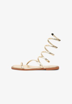 Flache Sandale mit goldfarbenen Metallriemen, darunter ein Spiralkleid, das sich um den Knöchel legt, und einer braunen flachen Sohle.