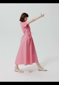 Femme en robe midi rose étirant les bras vers l'avant, debout sur une jambe étendue, portant des chaussures plates blanches sur un fond uni.