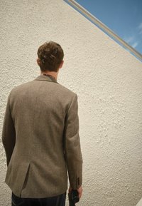 Mand i brun jakke holder en sort paraply, står vendt mod en struktureret hvid væg under en klar blå himmel.