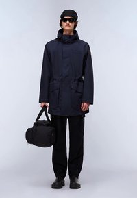 Granatowa wodoodporna kurtka z kapturem, elastycznym ściągaczem w talii oraz dwoma przednimi kieszeniami. Model trzyma czarną torbę, nosi ciemne spodnie i buty.