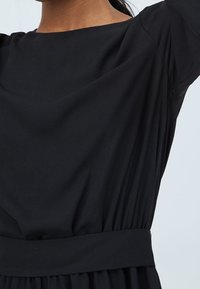 Svart klänning i mjukt material med rund halsringning, strukturerade ärmar, rynkad midja och ett brett bälte som betonar silhuetten.