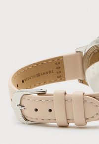 Beige nahkakellon hihna, jossa on valkoiset tikkaukset ja hopeinen solki, joka näyttää Tommy Hilfiger -brändin nimen ja sarjanumeron sisäpuolella.