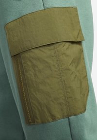 Světle zelená mikina z látky má velkou olivově zelenou nášivku se zipem a texturovaným povrchem po straně, což zdůrazňuje užitečnost a design.