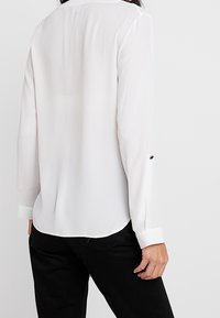 Bílá, lehká blůza s dlouhými rukávy, která má límec a knoflíkovou manžetu. Hladká textura s zakřiveným lemem a decentním detailem na zádech.