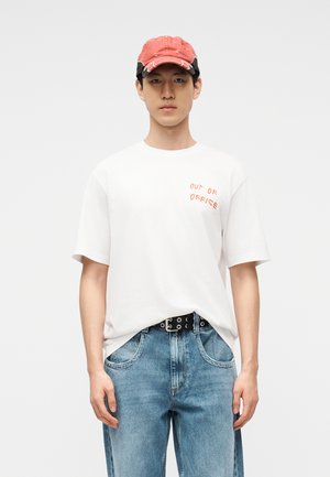 Nuori mies, jolla on kulunut punainen lippalakki, valkoinen t-paita, jossa lukee "OUT OF OFFICE", musta vyö ja siniset farkut, seisoo yksiväristä taustaa vasten.