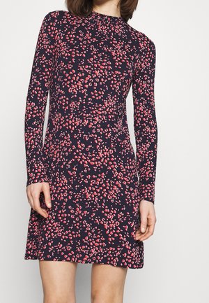 Mujer con un vestido azul marino de manga larga y largo hasta la rodilla, con un patrón abstracto de manchas rosas y rojas, sobre un fondo liso.