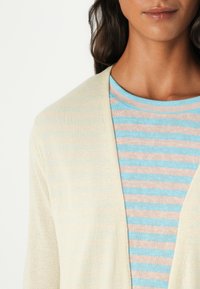 Cardigan beige clair en tissu doux, avec un devant ouvert et des manches longues, superposé à une chemise rayée bleue et rose.