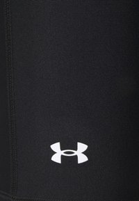 Czarne legginsy sportowe wykonane z gładkiego, elastycznego materiału, z widocznymi szwami i kontrastowym białym logo w dolnej części.