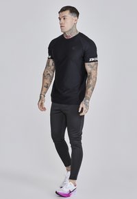 Fekete atlétikai póló rövid ujjakkal és SIKSilk logóval a mandzsettákon, fekete, testhez simuló sportnadrággal és fehér-lila futócipővel párosítva.