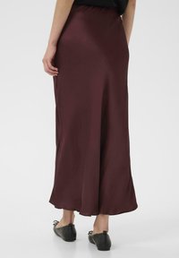 Burgunderfarbener Satin-Maxi-Rock mit elastischem Bund, glatter Textur und weichem Fall, kombiniert mit schwarzen flachen Schuhen, von hinten aufgenommen.