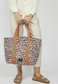 Osoba w jasnobeżowym stroju trzyma dużą torbę w panterkę z pomarańczowymi i zielonymi paskami, nosi beżowe sandały z klamrą.
