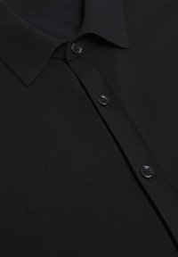 Musta ribbineuloksesta valmistettu poolopaita, jossa on terävä kaulus, kolme tumman sävyistä nappia ja sileä pinta.