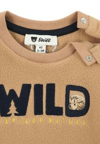Detailní záběr na béžový fleecový body pro miminko s textem "WILD", grafikou ježka v písmenu "D" a žlutou výšivkou "Explore Nature".