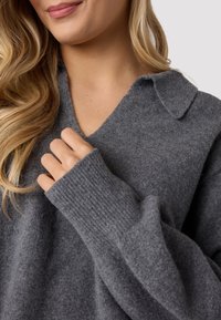 Maglione grigio in lana con scollatura a V e colletto, dotato di polsini a costine. Tessuto morbido, con una mano che afferra delicatamente il tessuto.