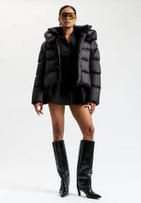Egy nő oversized fekete steppelt kabátot, rövid fekete ruhát, térd fölé érő fekete textúrált csizmát, sárgás árnyalatú napszemüveget és ezüst fülbevalót visel.