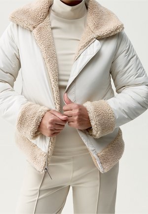 Person, der lynlås en cremefarvet vinterjakke med sherpa-foring på krave, manchetter og kant, iført en beige rullekrave og bukser.