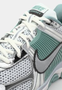 Nike Vomero 5 tenisice imaju gornji dio od mrežice s srebrnim i tirkiznim naglascima, reflektirajuće detalje, podstavljeni đon i bijele vezice.