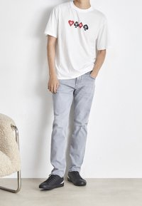 Maglietta bianca con grafica rossa e nera con la scritta "HUGO", jeans azzurri chiari e sneakers nere con accenti tricolore. Sfondo semplice.