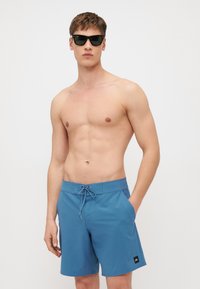 Blaue Badeshorts mit einem Zugbänderverschluss und Seitentaschen. Der Stoff wirkt glatt, leicht und schnelltrocknend. Schwarzes Logo-Patch am Saum.