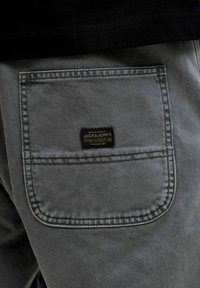 Κοντινό πλάνο στην πίσω τσέπη γκρι τζιν παντελονιού με μια μαύρη και χρυσή ετικέτα της μάρκας Jack & Jones ραμμένη στο κέντρο.