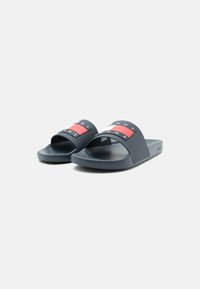 Fekete slip-on szandálpár piros, fehér és kék téglalap alakú logóval az elülső pánton, fehér háttérrel.