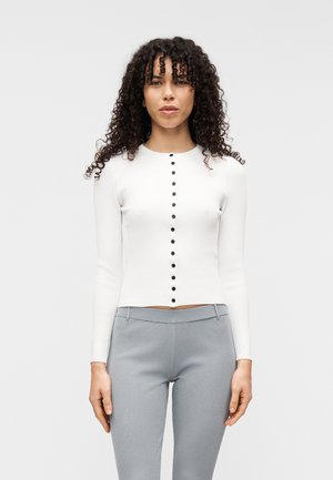 Cardigan blanc à côtes avec manches longues et encolure ronde, doté d'une rangée de boutons noirs sur le devant. Associé à un pantalon gris clair texturé.