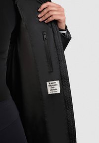 Schwarze Jacke mit glatter Textur, ausgestattet mit einer Reißverschlusstasche innen und einem Etikett mit der Aufschrift „Eine warme Umarmung aus Stoff.“