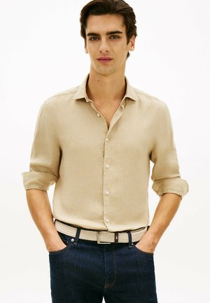 Młody mężczyzna o ciemnych włosach, ubrany w beżową koszulę na guziki z podwiniętymi rękawami, ciemne dżinsy i pleciony beżowy pasek, ręce w kieszeniach.